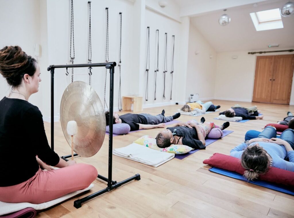 A practitioner plays a gong during a sound bath session as people lie on their backs with closed eyes.