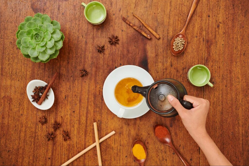 Woman pouring tea into a cup next to herbs, spices, and plants.