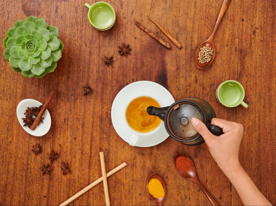Woman pouring tea into a cup next to herbs, spices, and plants.