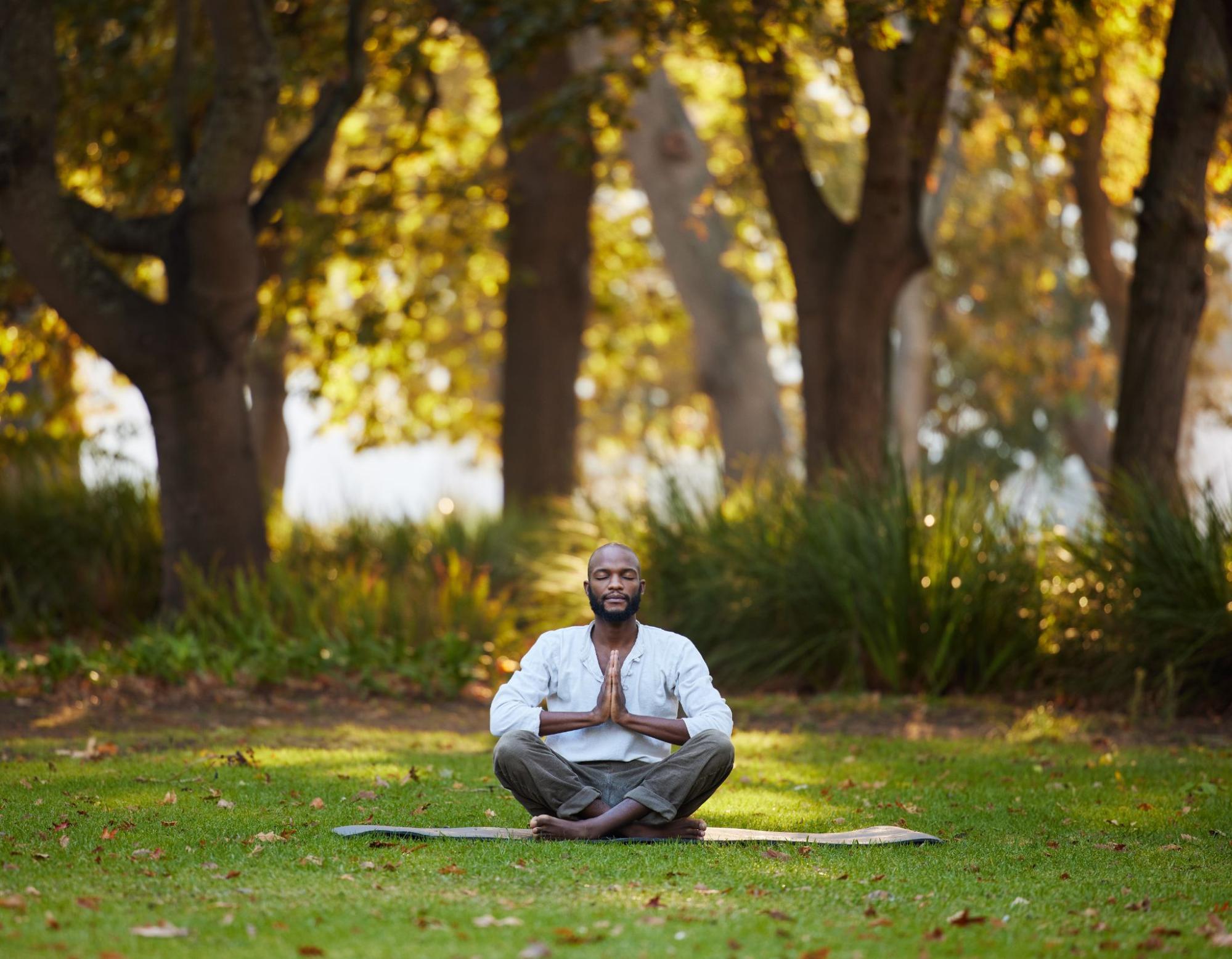 Full-body shot of a young man meditating outdoors in a park.