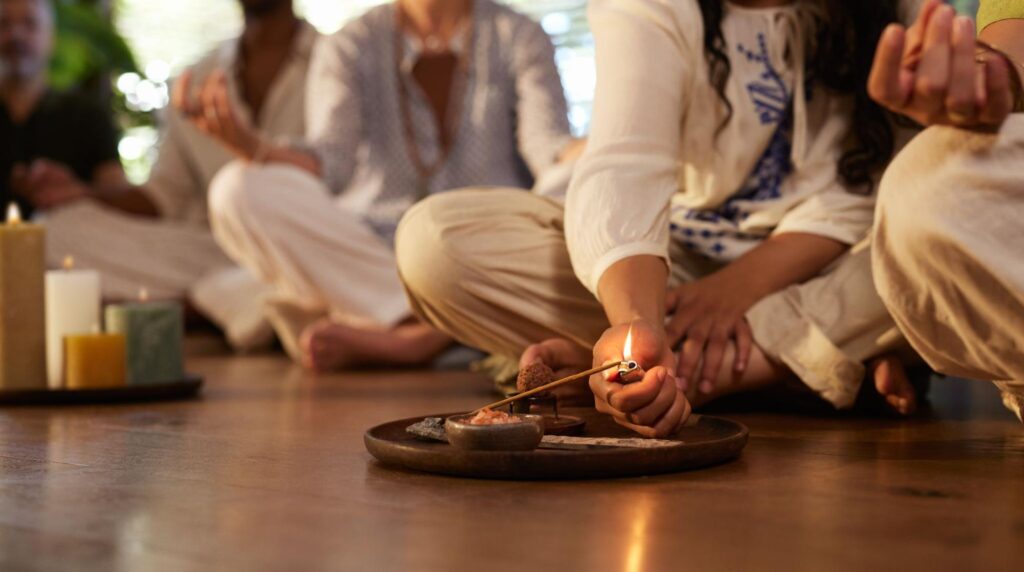 A group of people sitting on the floor posing in a spiritual practice while a woman lights incense.