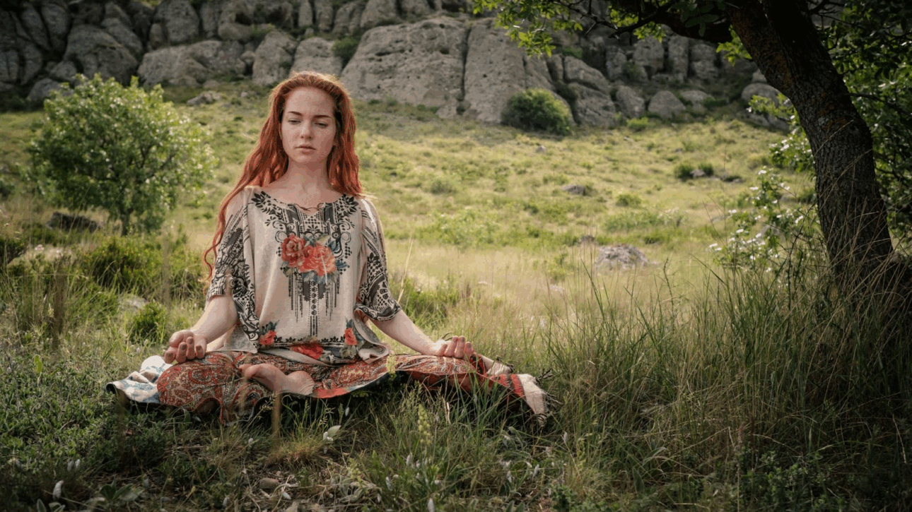 A young woman meditates under a tree in a large park.