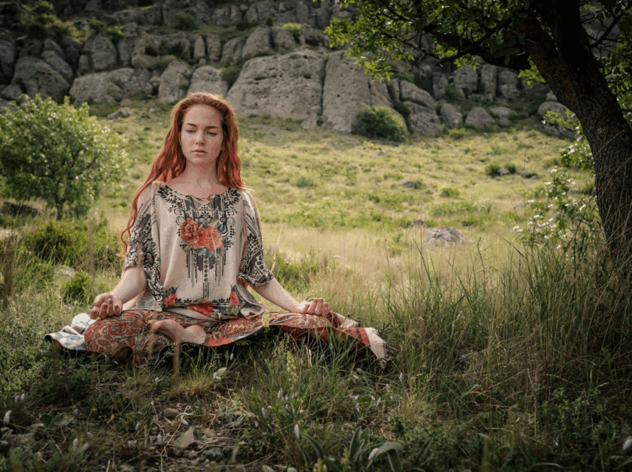 A young woman meditates under a tree in a large park.