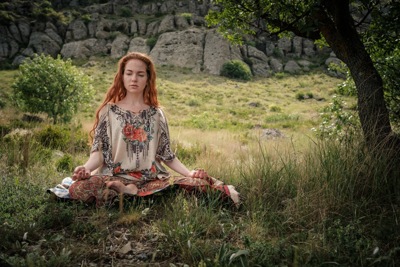 A young woman meditates under a tree in a large park.