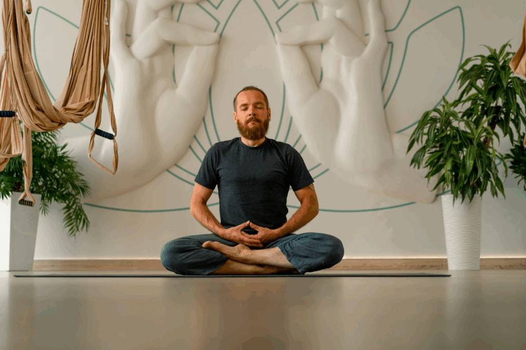 A man meditates in a studio with meditation art in the background.