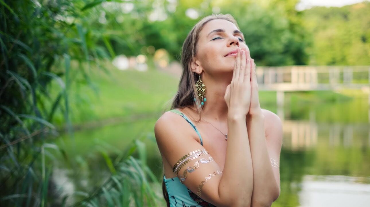 Woman standing in green nature at sunset, praying quietly and embracing spiritual reflection.