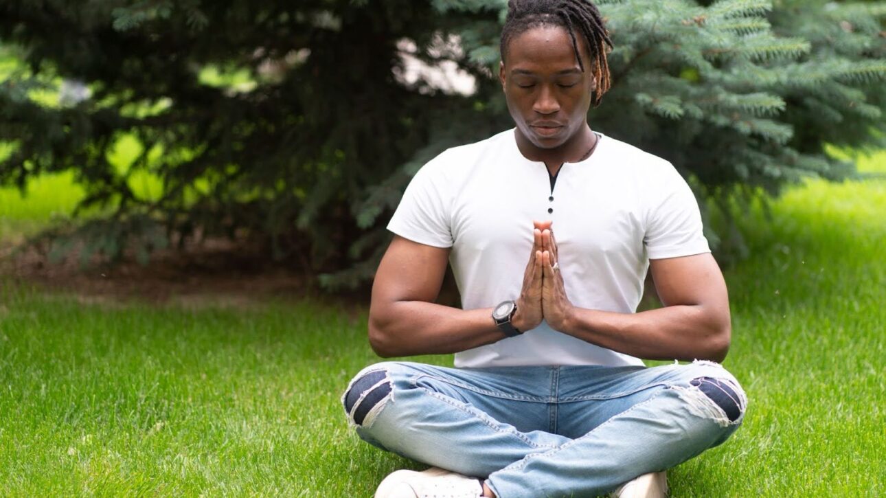 A man meditating in a seated yoga pose on a green park lawn.