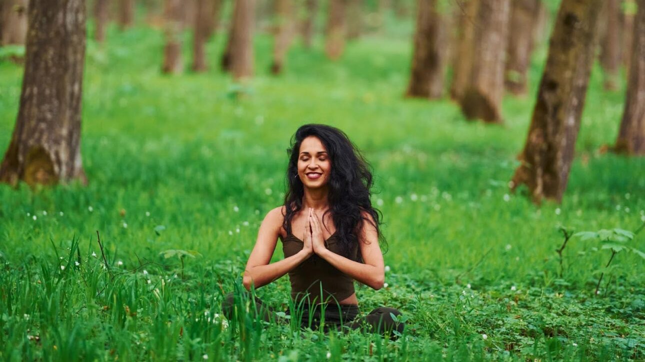 A woman sits in a forest, meditating with her hands pressed together.