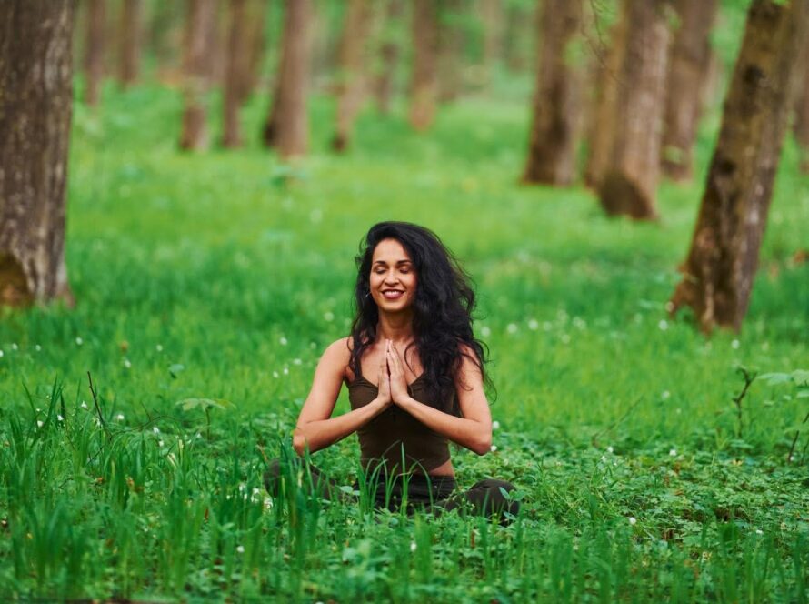 A woman sits in a forest, meditating with her hands pressed together.