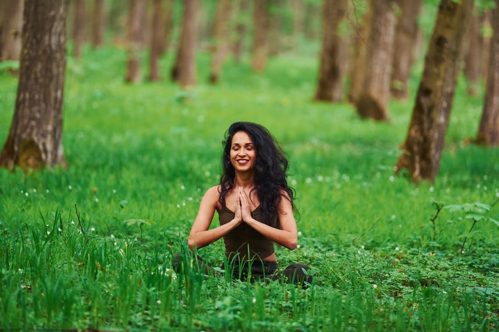 A woman sits in a forest, meditating with her hands pressed together.