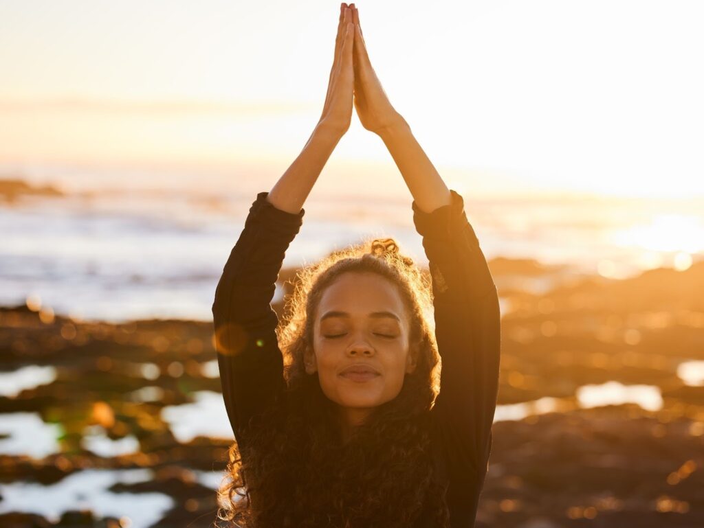 A young woman does yoga on the beach by reaching above her head.