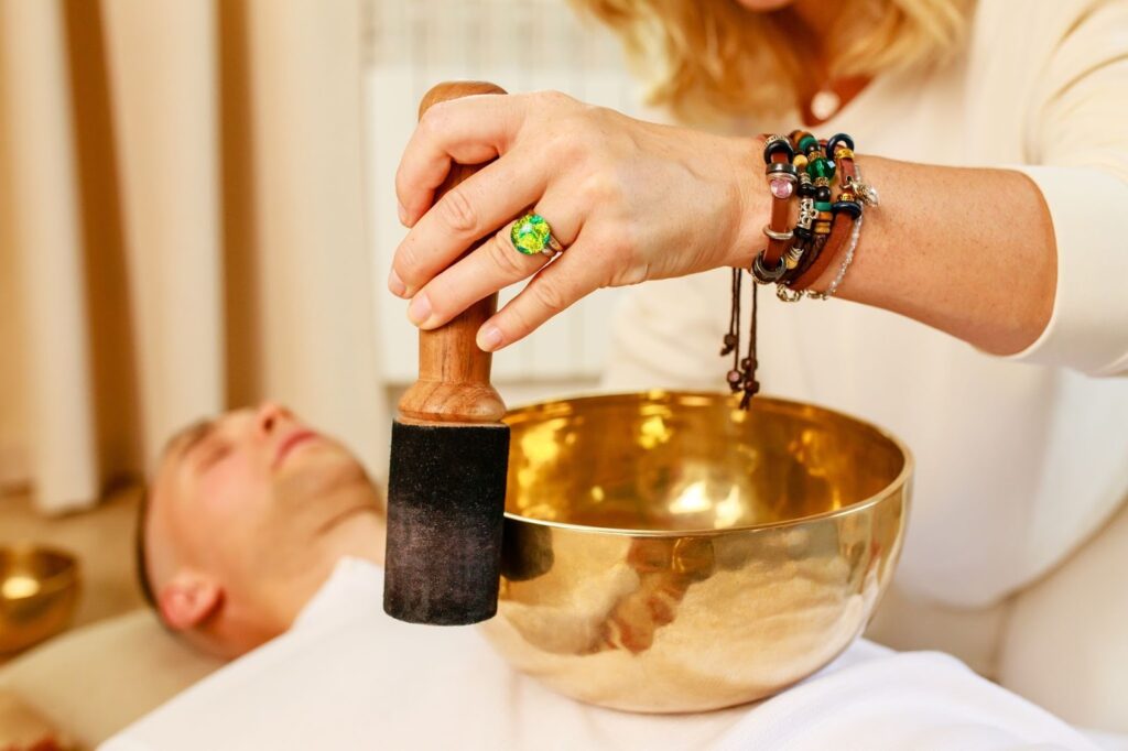 A woman plays a singing bowl on a mans chest while he lies down for a sound bath.