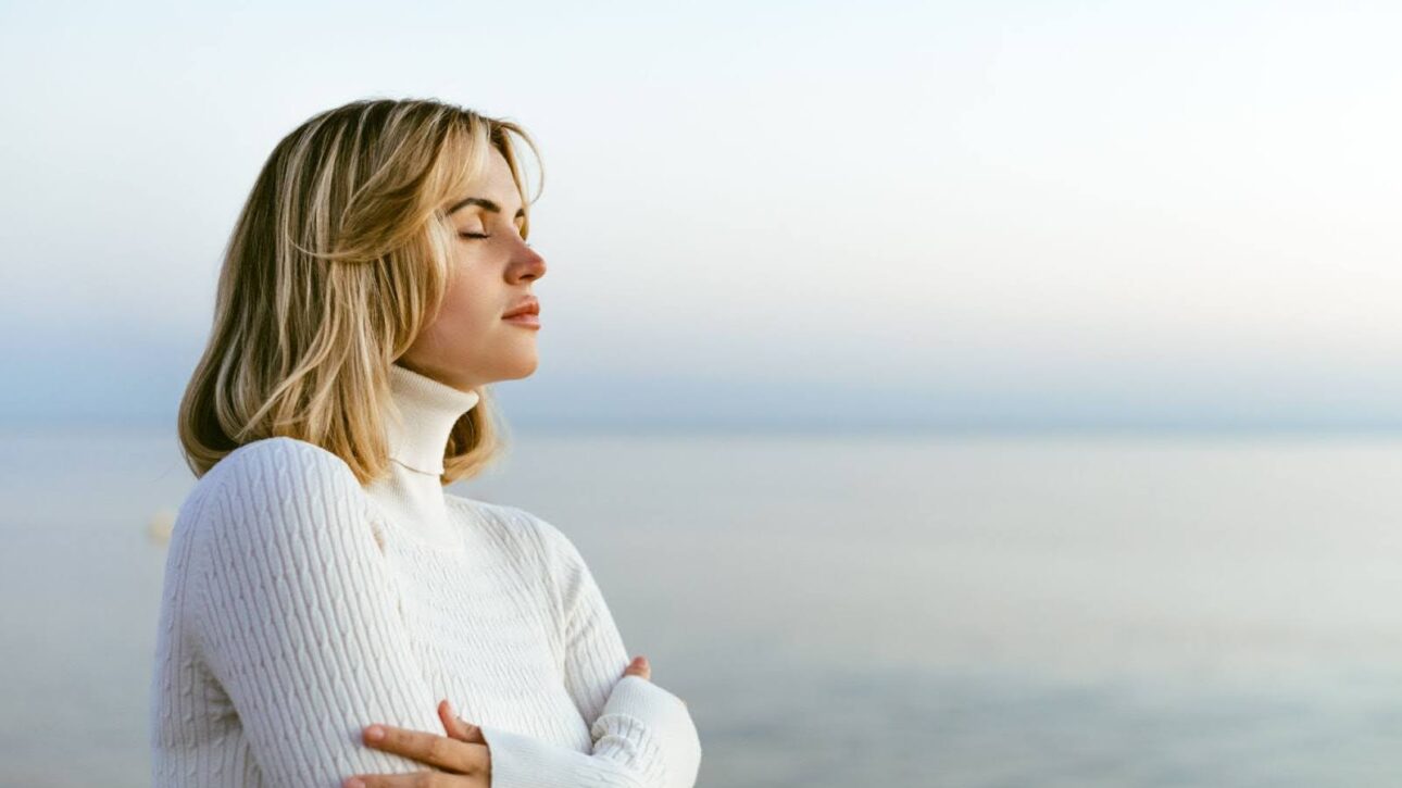 Beautiful woman in a sweater practicing mindful meditation on a calm seaside shoreline.