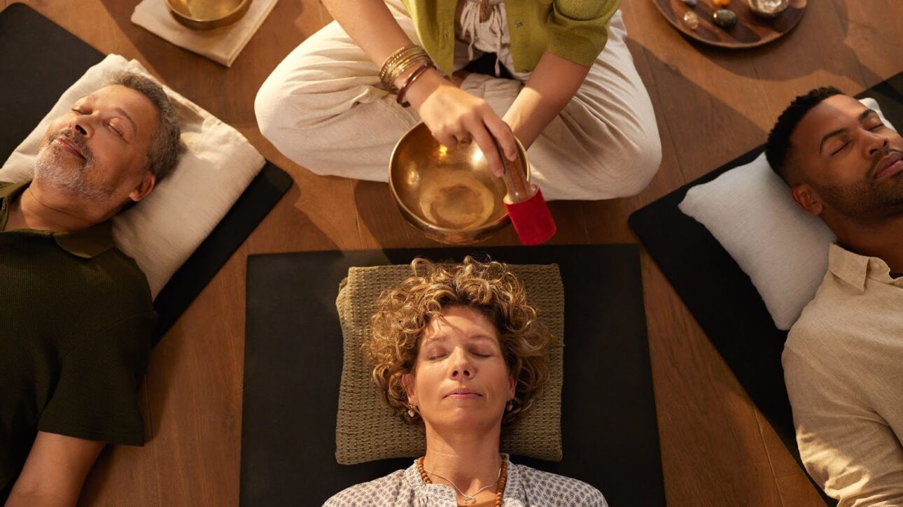 Overhead view of a sound healing session with three individuals lying down while a practitioner creates sound from a golden singing bowl.