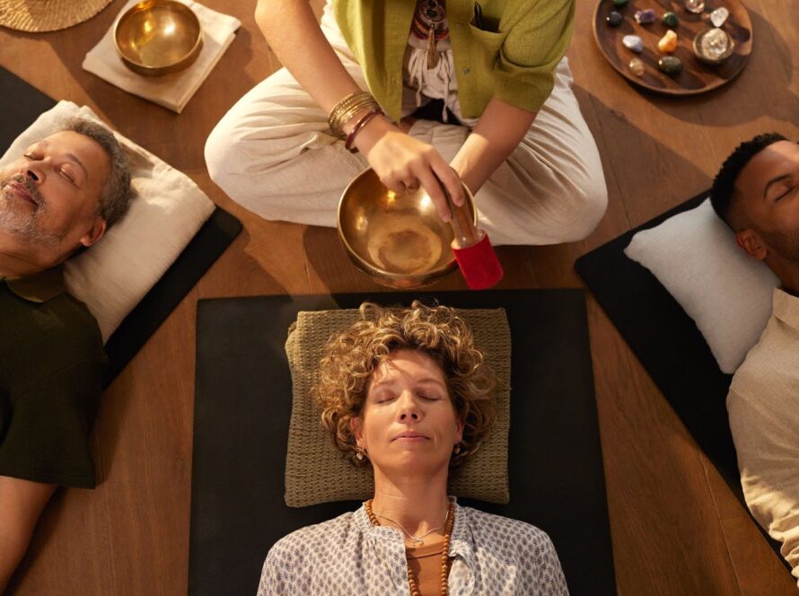 Overhead view of a sound healing session with three individuals lying down while a practitioner creates sound from a golden singing bowl.