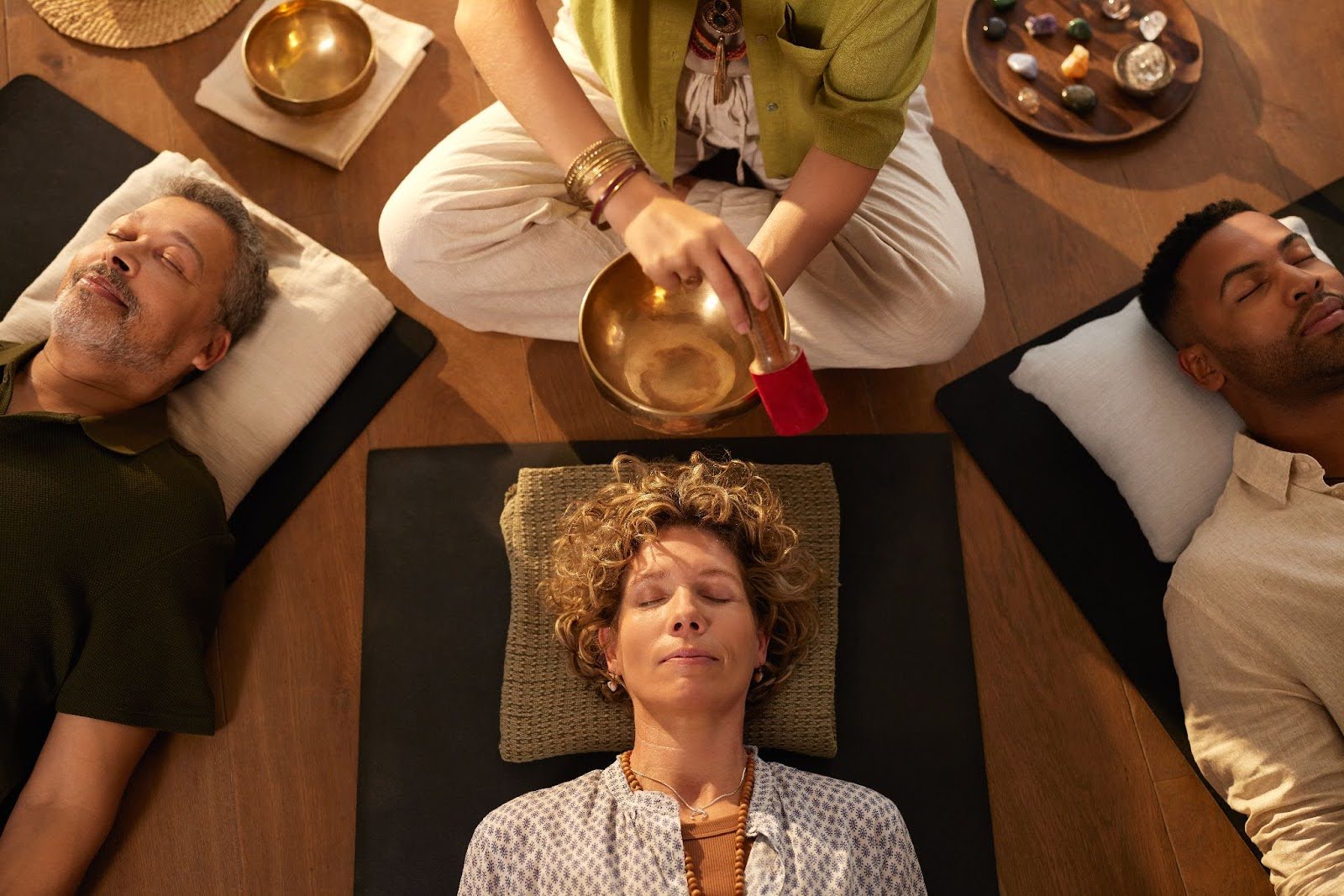 Overhead view of a sound healing session with three individuals lying down while a practitioner creates sound from a golden singing bowl.