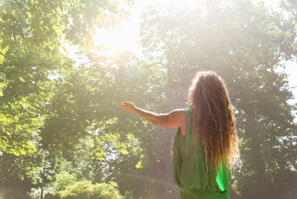 Young woman in a green top standing in nature with arms outstretched, symbolizing spiritual energy and connection.