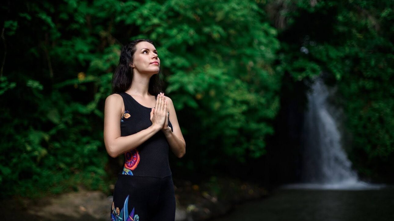 Woman on a spiritual journey with her hands folded in prayer next to water and foliage in the background.