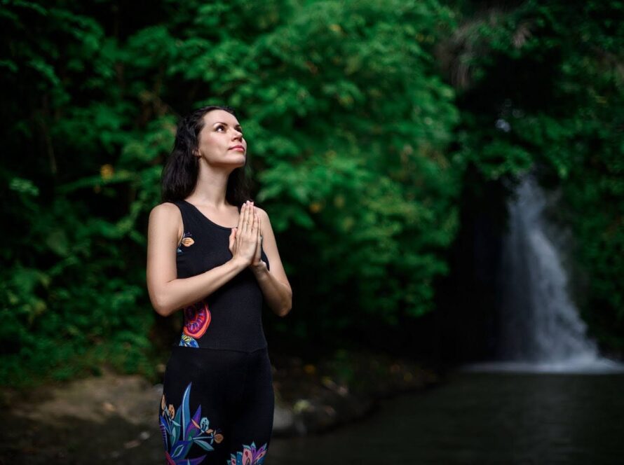 Woman on a spiritual journey with her hands folded in prayer next to water and foliage in the background.