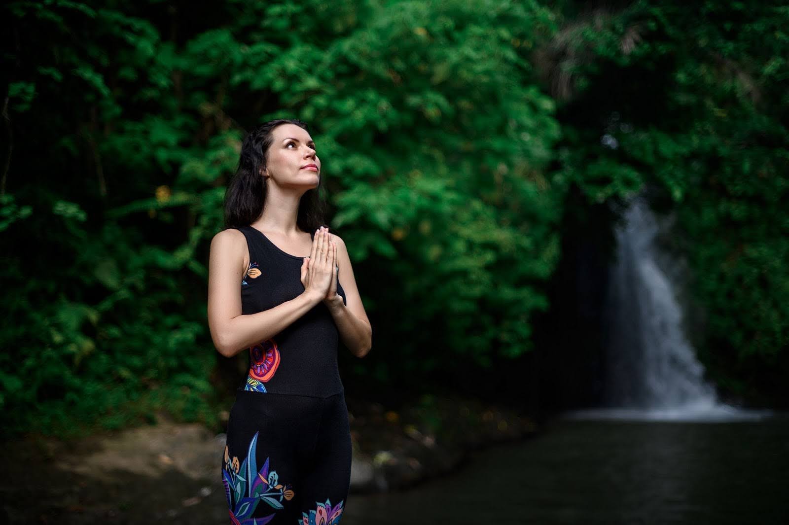 Woman on a spiritual journey with her hands folded in prayer next to water and foliage in the background.