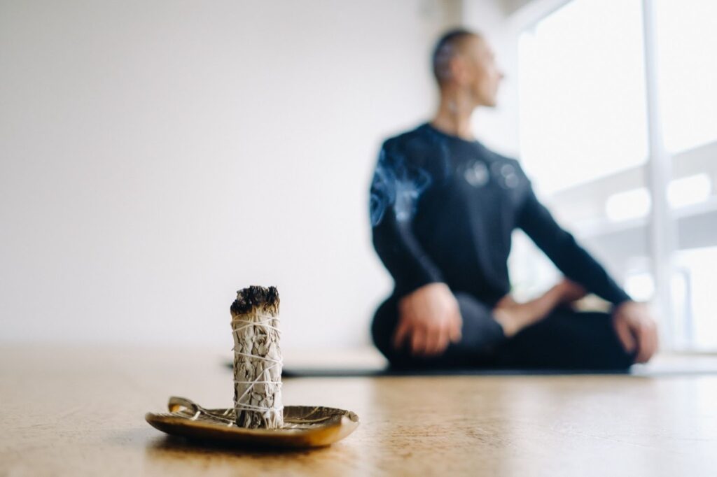 Male athlete in black sportswear meditating in lotus position near incense, reflecting self-discipline and abstinence from training.