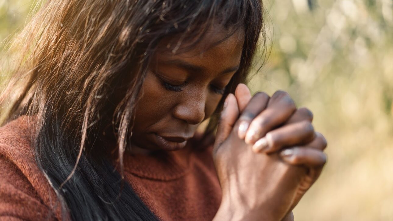 A Black woman standing outdoors in focused prayer, expressing faith and devotion