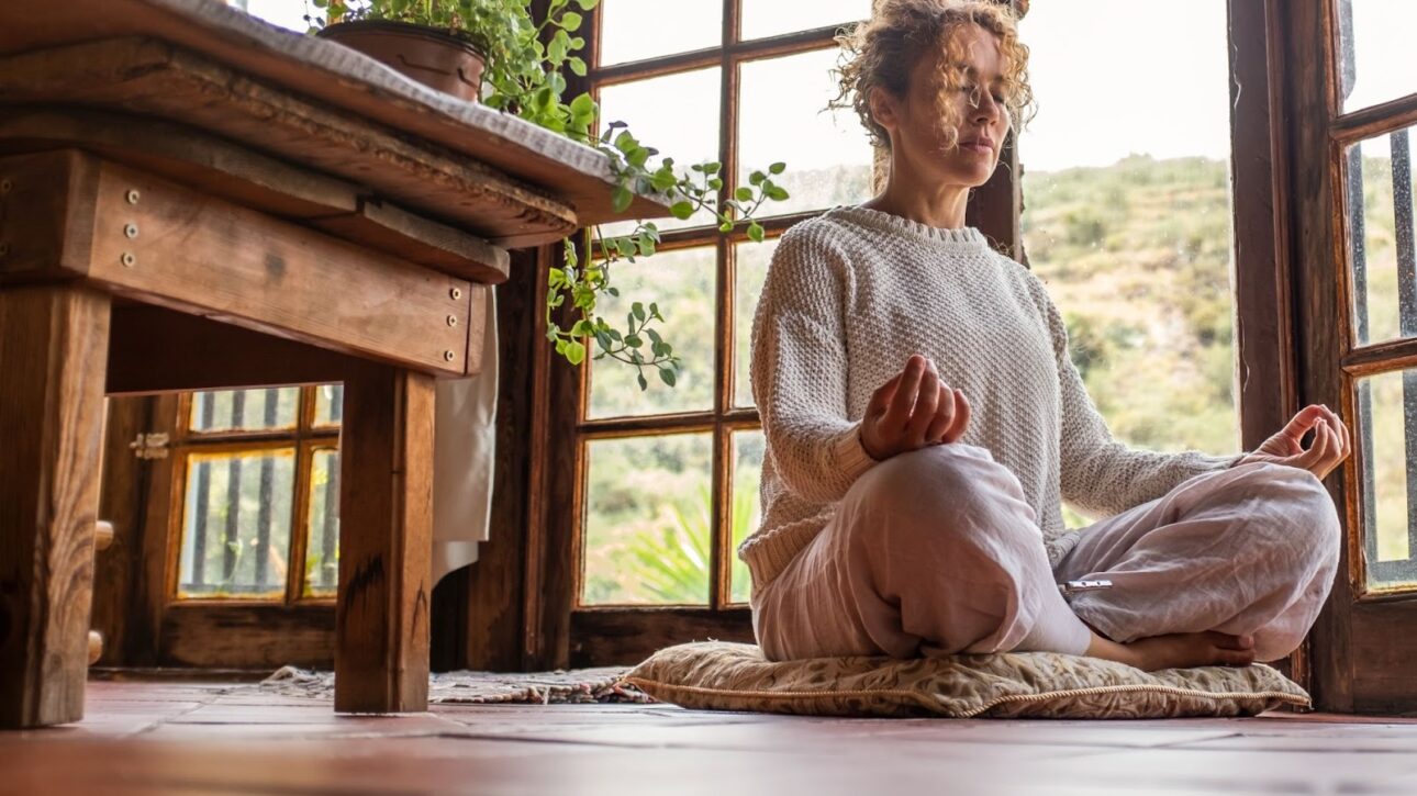 Woman with her eyes closed and her legs crossed next to a table and an open door. 