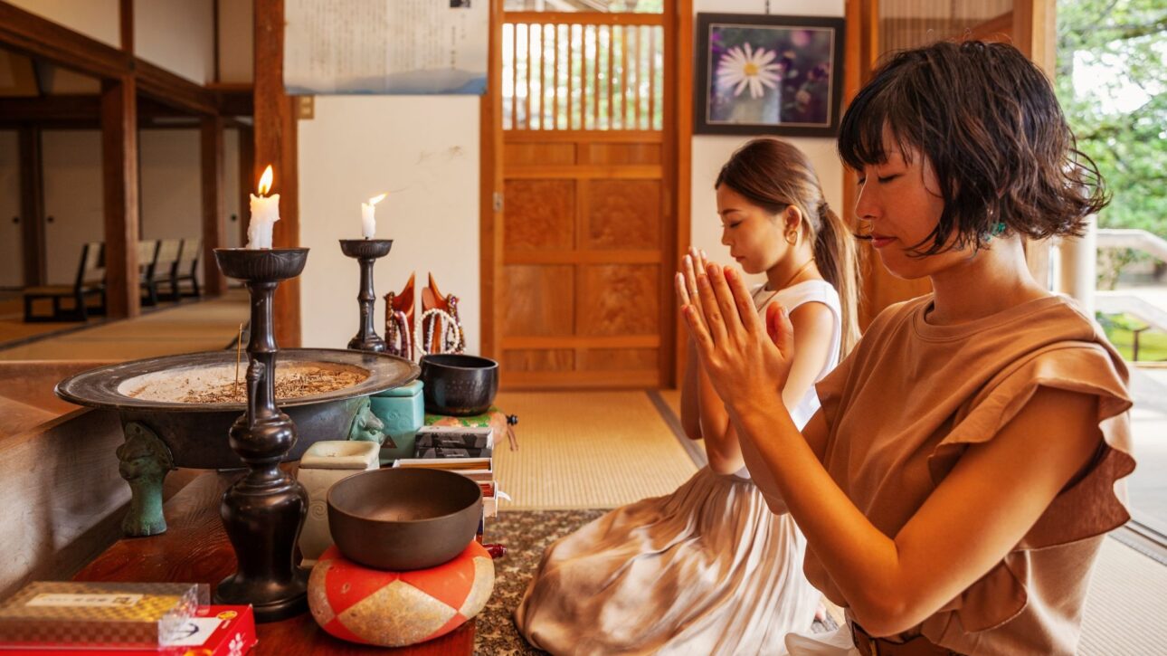 Two women kneel in prayer at a wooden shrine altar with lit candles and offerings.
