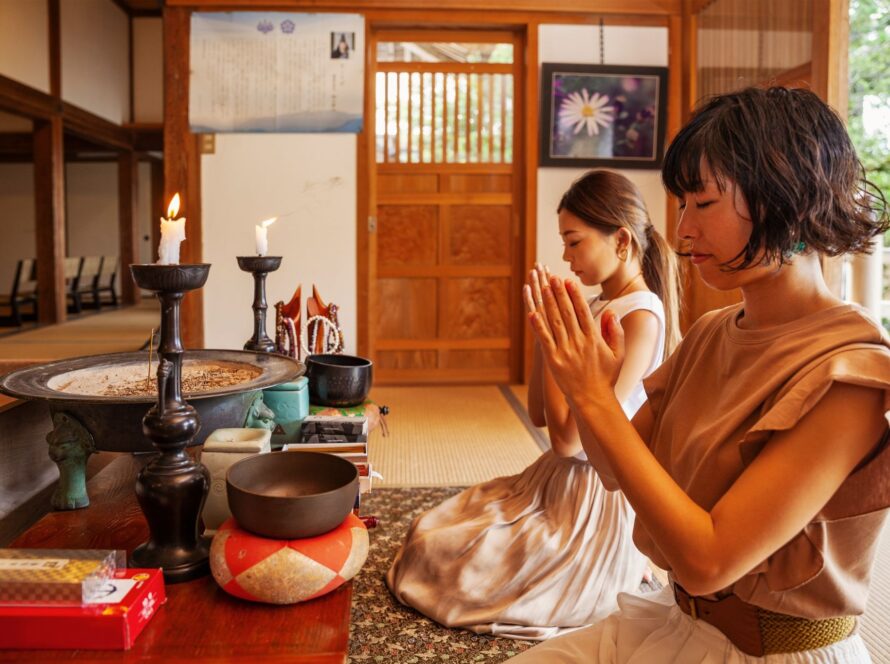 Two women kneel in prayer at a wooden shrine altar with lit candles and offerings.