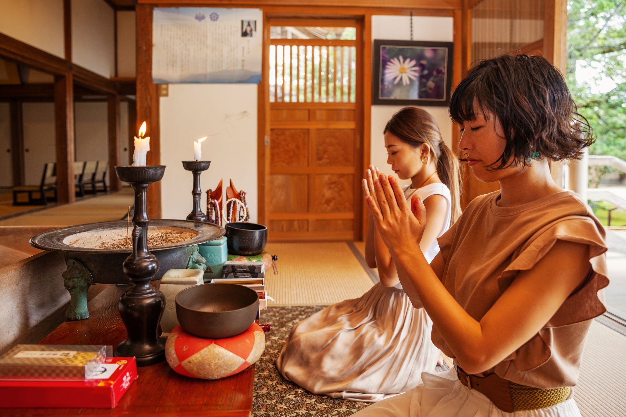 Two women kneel in prayer at a wooden shrine altar with lit candles and offerings.