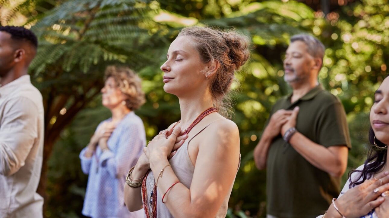 Diverse community gathered outdoors, standing in reflective meditation amid lush greenery