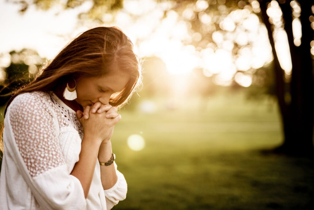 A woman praying in a green meadow at sunset, standing in soft golden light with her head bowed down